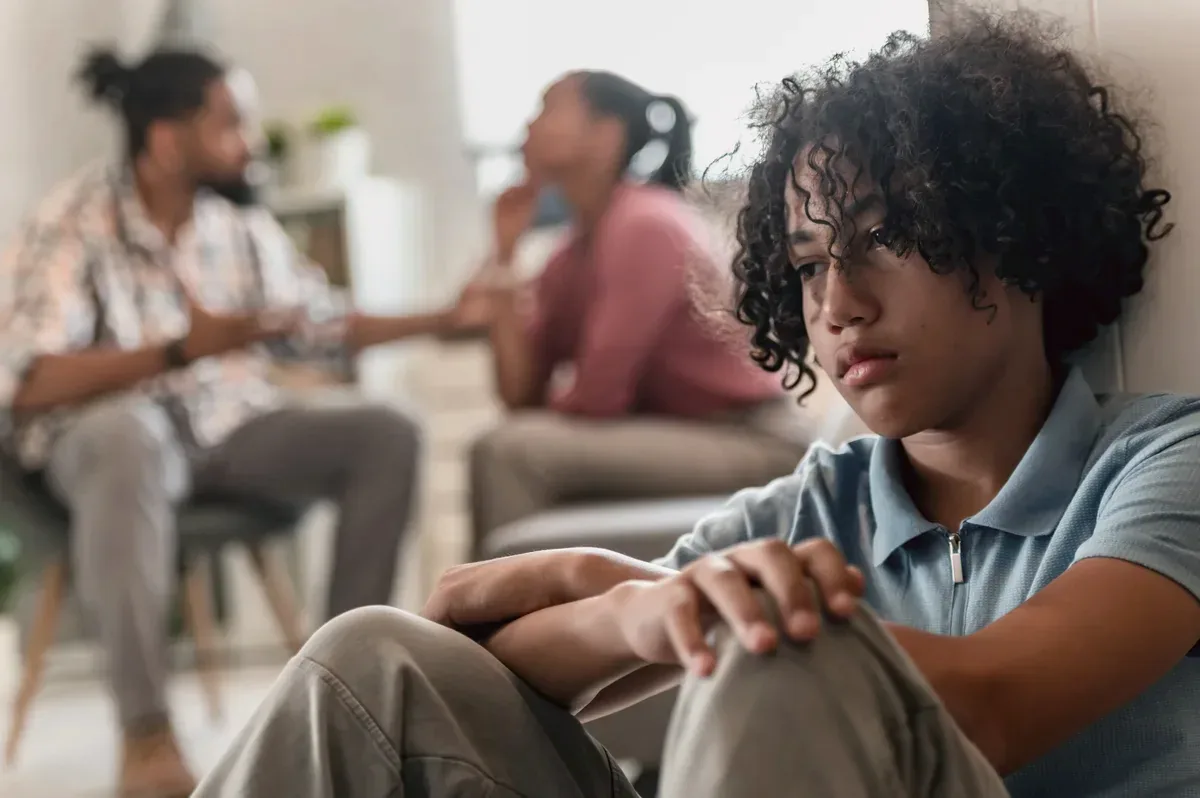 A young person sitting pensively while their parents talk in the background