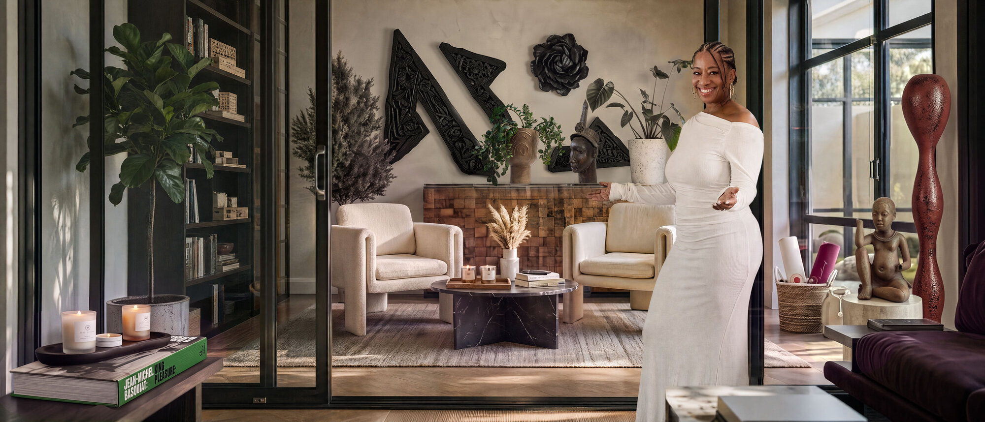 Lisane Basquiat in a cream off-the-shoulder gown in a sunlit living room, smiling and gesturing toward two armchairs, carved wall art, and shelves of books
