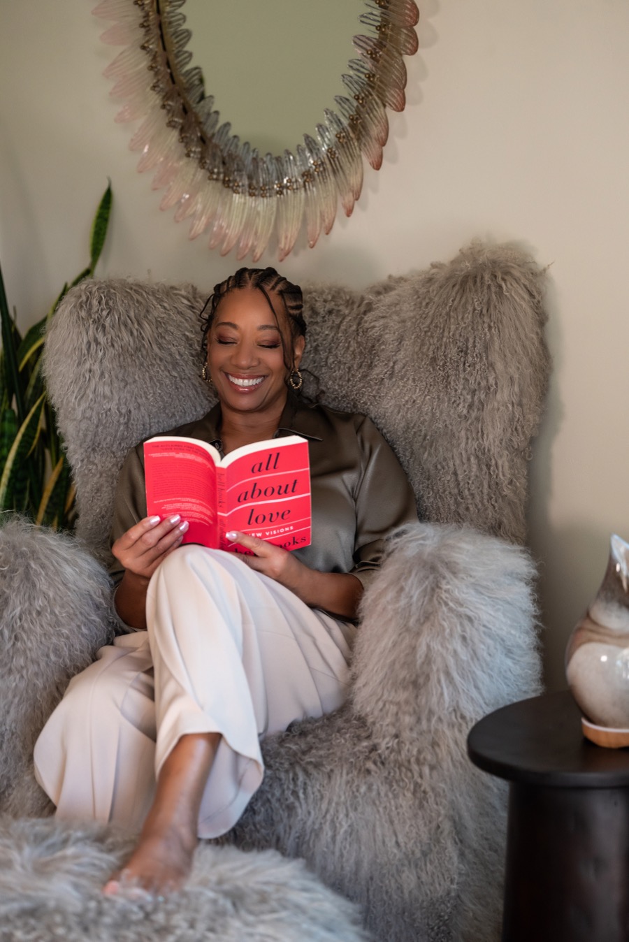 Lisane Basquiat smiling, reading All About Love by bell hooks in a cozy chair