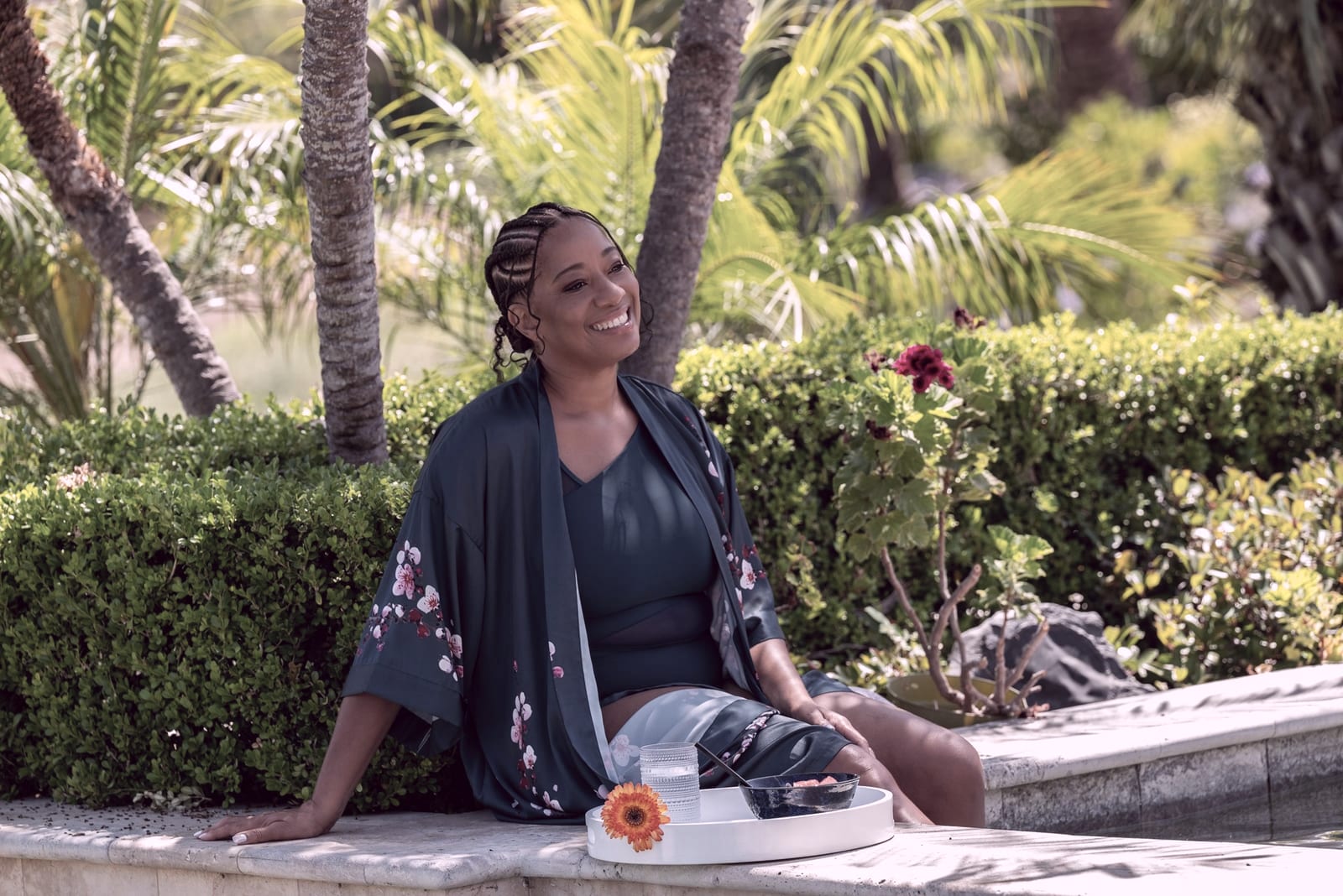 Lisane Basquiat laughing, seated on the edge of a garden pool in a teal floral kimono, palms and a flowering hedge behind her