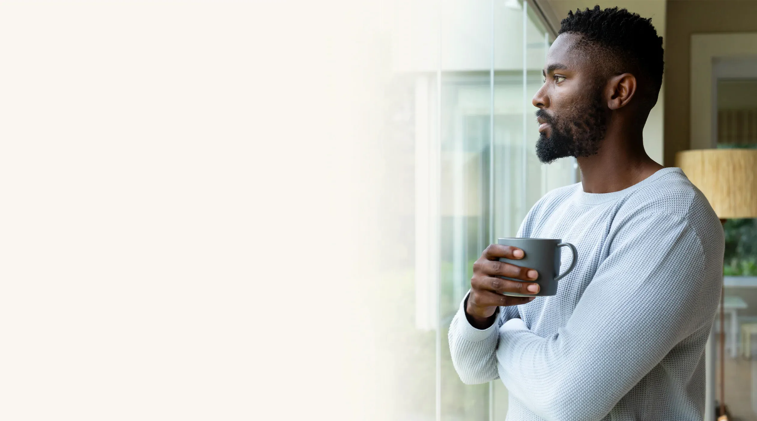 A man holding a mug, looking out a window, standing at ease