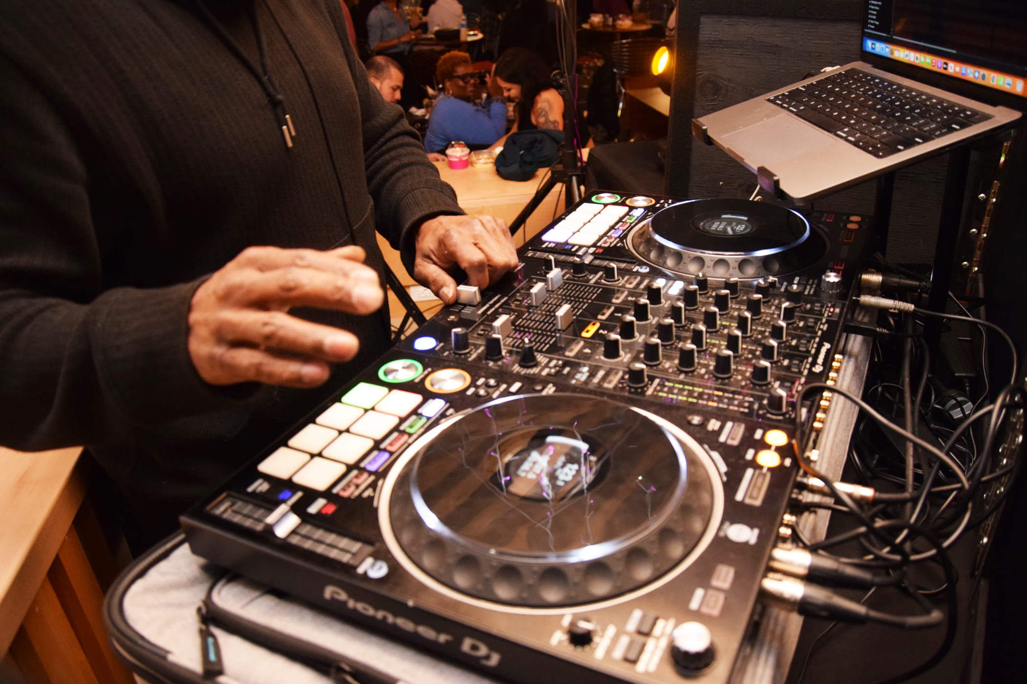 A DJ mixing at a Soulful Socials gathering, guests talking around tables behind the decks